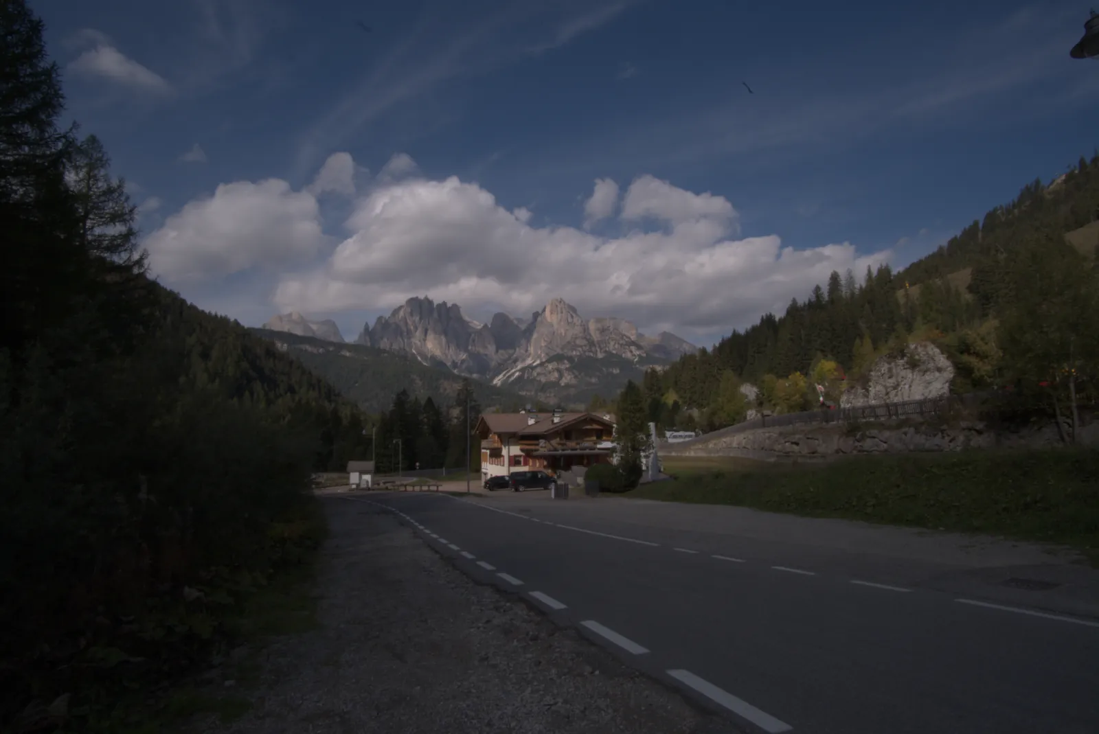 Strada di montagna verso le guglie delle Dolomiti sotto grandi nuvole, così com'esce dal RAW: bosco e strada scuri, cielo spento